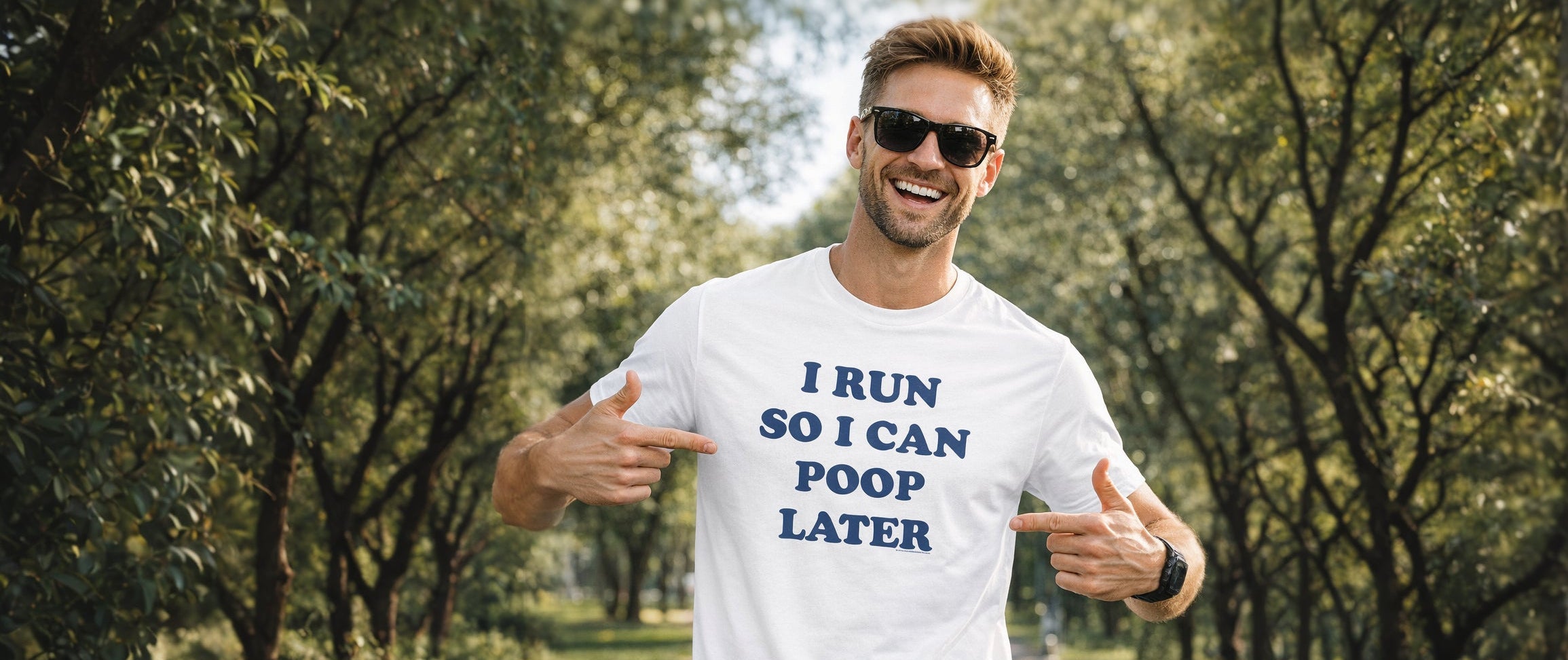 A man wearing a white graphic t-shirt with the text 'I RUN SO I CAN POOP LATER' in blue, standing on a tree-lined path and pointing at the shirt.
