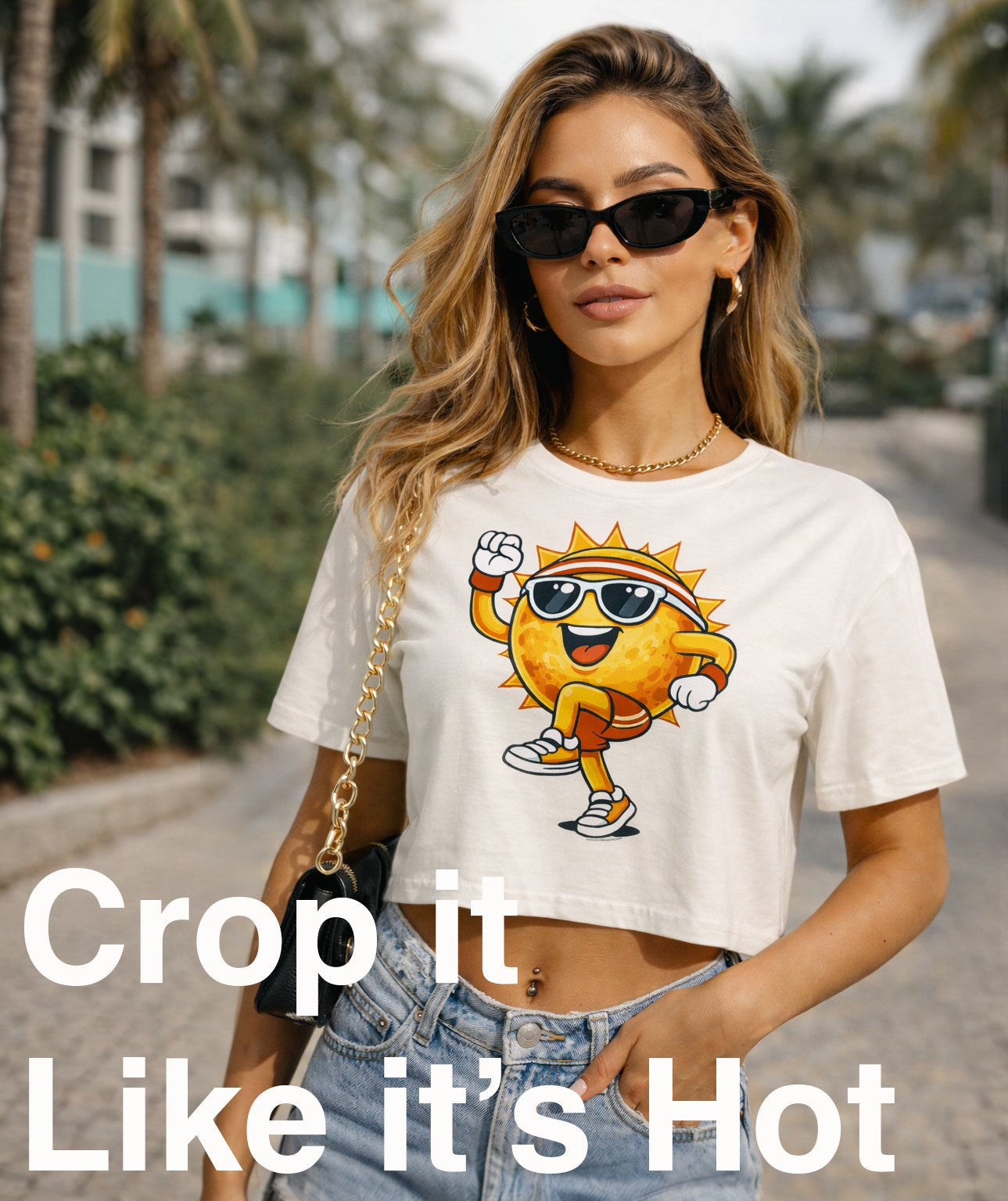 A woman wearing a white cropped graphic T-shirt with a cartoon sun character and light blue jeans, standing outdoors on a paved walkway with palm trees in the background. Large text at the bottom reads 'Crop it Like it's Hot'.